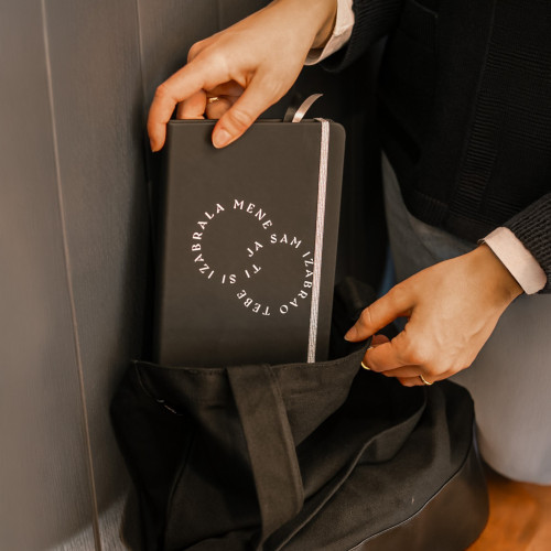 Crni premium rokovnik tvrdih korica &ldquo;Ti si izabrala mene, ja sam izabrao tebe&rdquo;, koji djevojka pažljivo izvlači iz torbe iz Dino Merlin kolekcije.