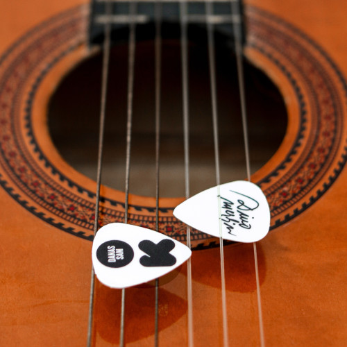 Bijele trzalice za gitaru sa crnim printom "Danas sam OK" inspirisane stihom iz pjesme Dine Merlina "Danas sam OK" s albuma ''Nešto lijepo treba da se desi'' na žicama od smeđe gitare.