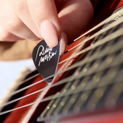 Crna trzalica za gitaru sa bijelim potpisom "Dino Merlin" u ruci djevojke koja prelazi preko žica po crvenoj gitari.