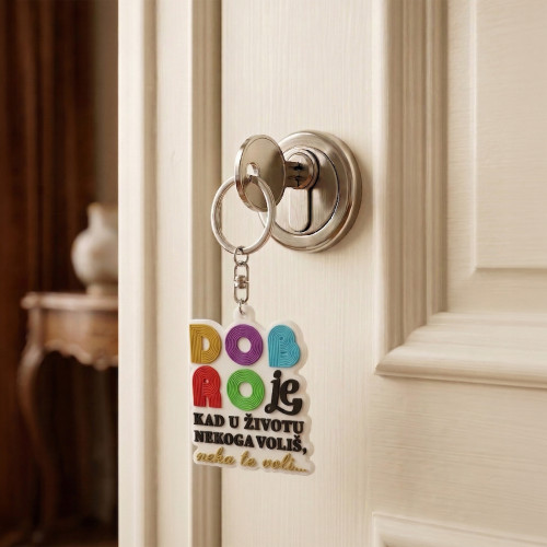 Bijeli privjesak za ključeve sa &scaron;arenim slovim"Dobro je kad u životu nekoga voli&scaron;, neko te voli" inspirisan iz pjesme Dine Merlina "Da &scaron;uti&scaron;" u ključaonici bijelih vrata.