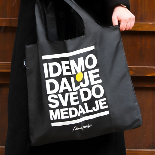 Crni ceker  (torba) za shopping sa futrolom sa natpisom "Idemo dalje sve do medalje" u ruci djevojke u kaputu na smeđoj pozadini.