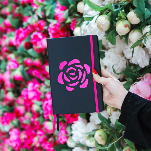 Crni rokovnik tvrdih korica "Neka bude Đula" s pink ružom u ruci djevojke na &scaron;arenom cvjetnom zidu.