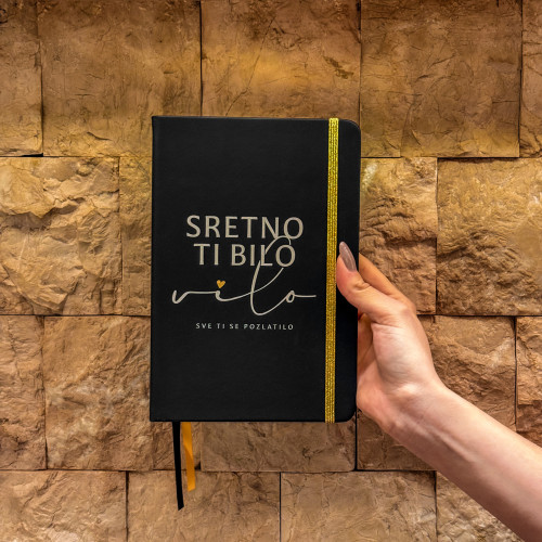 Crni premium rokovnik &ldquo;Sretno ti bilo, vilo, sve ti se pozlatilo&rdquo; inspirisan stihom Dine Merlina, prikazan uz &scaron;oljicu kafe na drvenom stolu.