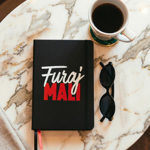 Crni premium rokovnik tvrdih korica &ldquo;Furaj mali&rdquo; na bijelom stolu s kafom i crnim naočalama pored.