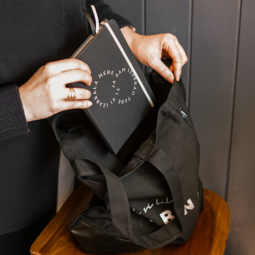 Crni premium rokovnik tvrdih korica &ldquo;Ti si izabrala mene, ja sam izabrao tebe&rdquo;, koji djevojka pažljivo izvlači iz torbe iz Dino Merlin kolekcije.