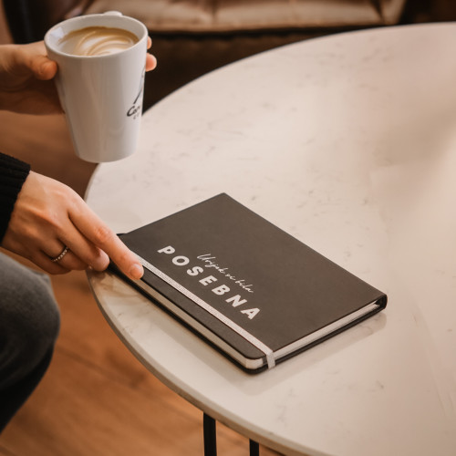 Crni premium rokovnik "Uvijek si bila posebna" s rukom djevojke koja drži &scaron;olju s kafom na bijelom stolu. 