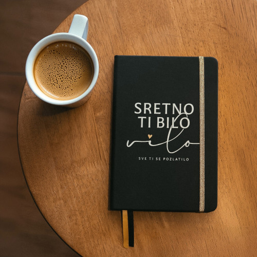 Crni premium rokovnik &ldquo;Sretno ti bilo, vilo, sve ti se pozlatilo&rdquo; inspirisan stihom Dine Merlina, prikazan uz &scaron;oljicu kafe na drvenom stolu.