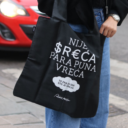 Crni ceker/torba sa stihom 'Nije sreća para puna vreća' u ruci djevojke u bijelom džemperu. 